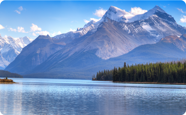A serene lake with calm water reflects tall evergreen trees and towering snow-capped mountains under a clear blue sky with scattered clouds.